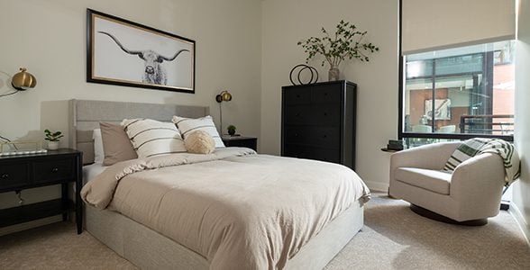 Bedroom in a Ballantyne apartment with a bed, chair, dresser, and large windows